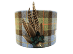 A cylindrical lampshade made of Harris Tweed with a plaid pattern and decorated with real pheasant feathers attached to the top.