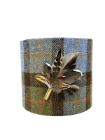 A cylindrical lampshade made of Harris Tweed with a plaid pattern and decorated with real pheasant feathers attached to the top.