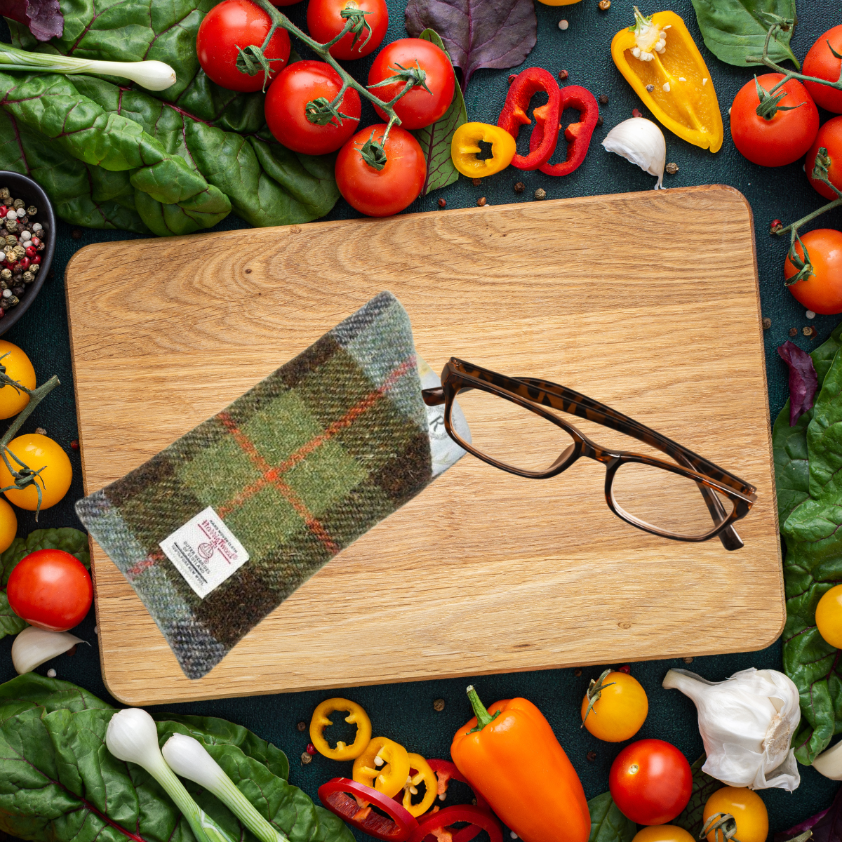 A glasses case made of Harris Tweed is displayed on a wooden board, surrounded by an array of colorful vegetables. The case is partially opened to reveal a pair of glasses resting inside.